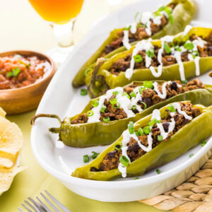 Längs aufgeschnittene grüne Chilischoten mit Hackfleisch-Bohnen-Füllung auf einer weißen ovalen Platte, mit dünnem Sauerrahm-Zickzack und Frühlingszwiebelringen, daneben Salsa, Tortilla-Chips und Limette