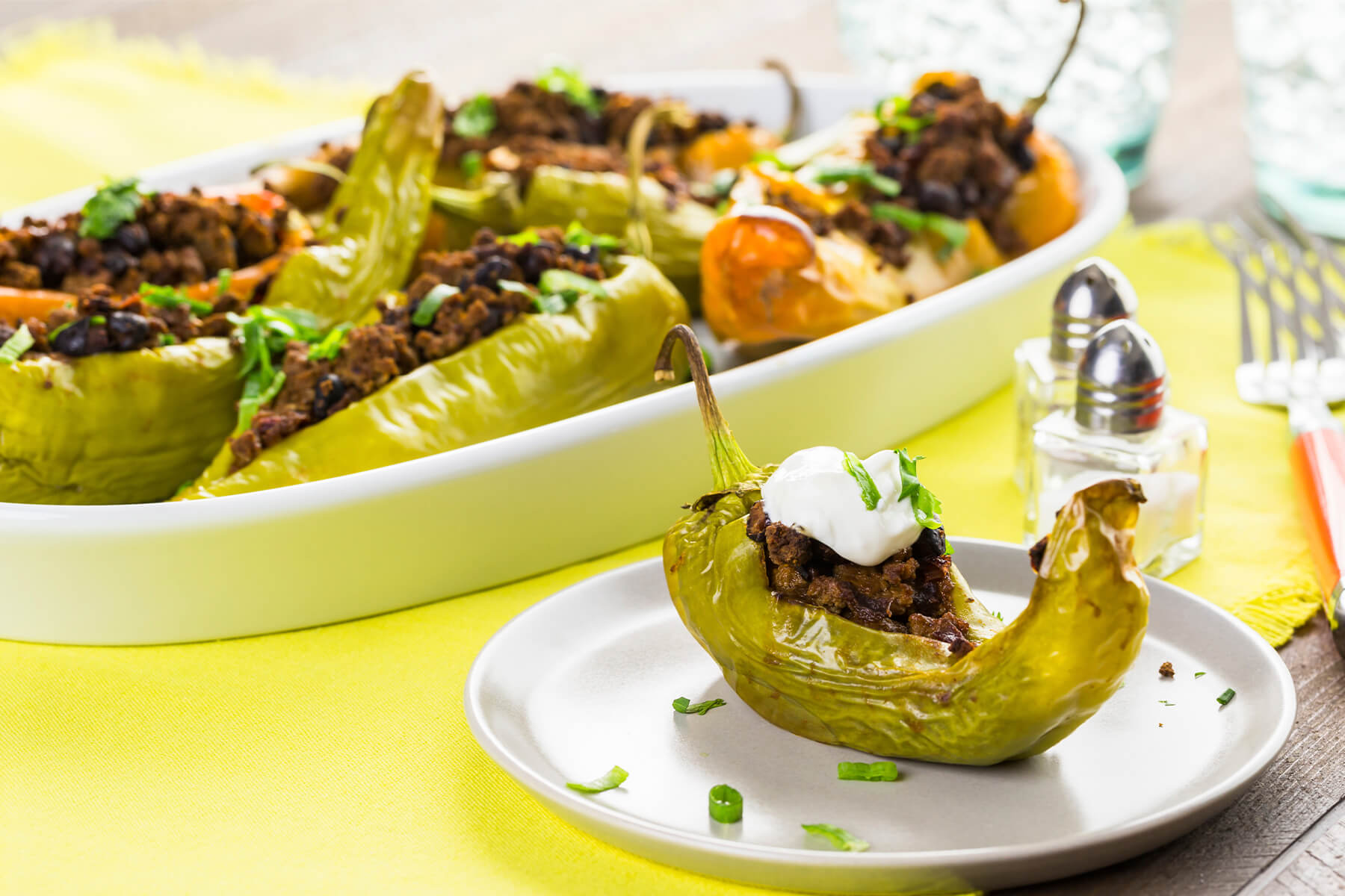 Gefüllte Chilischoten dicht nebeneinander in einer weißen Auflaufform, davor eine einzelne Schote auf einem Teller mit einem Klecks saurer Sahne und Korianderblättchen