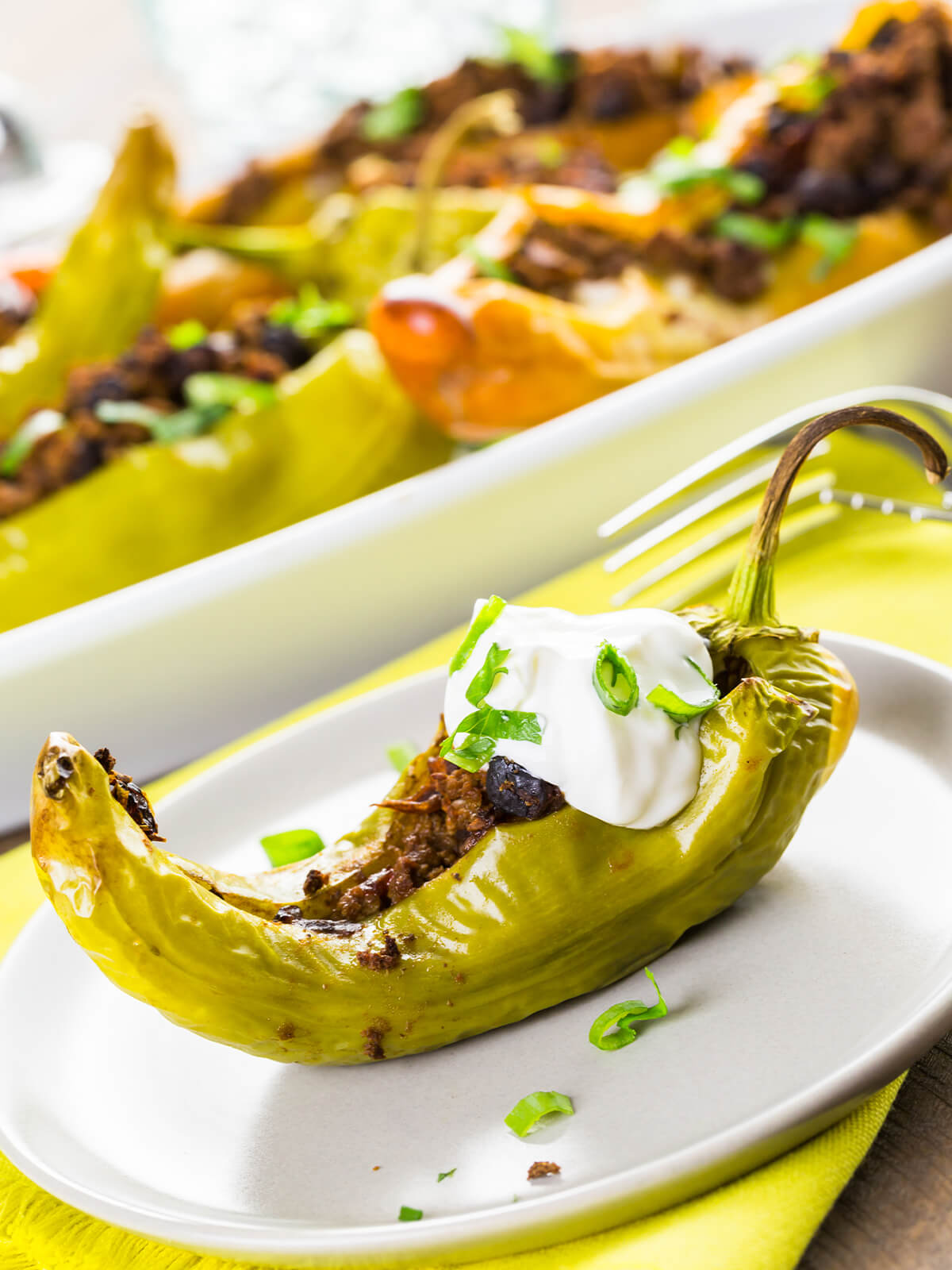 Einzelne gefüllte Chilischote mit Stiel auf einem weißen Teller, obenauf ein Klecks saure Sahne mit Frühlingszwiebelringen, dahinter die Auflaufform