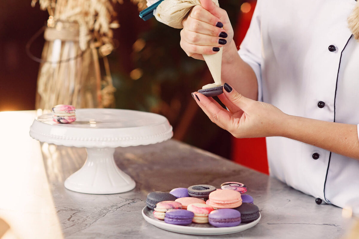 Aus einem Spritzbeutel ohne Tülle wird helle Creme auf eine anthrazitfarbene Macaronschale gespritzt, die in der Hand gehalten wird