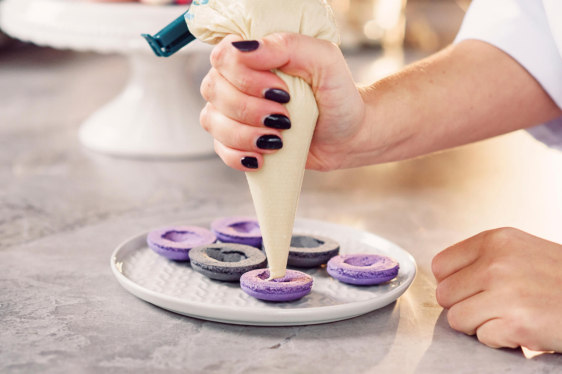 Aus einem Spritzbeutel ohne Tülle wird helle Creme in die Mulde einer violetten Macaronschale gespritzt