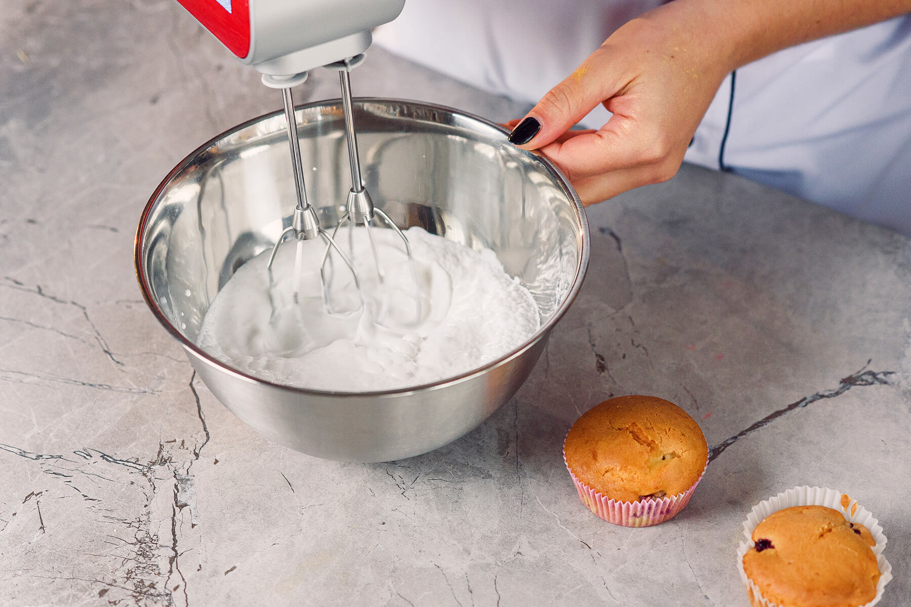 Eine Edelstahlschüssel mit weißer aufgeschlagener Crememasse steht neben zwei fertig gebackenen Muffins auf einer Marmorplatte