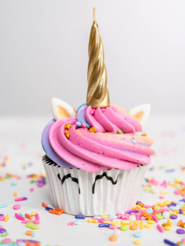 Einhorn-Cupcake mit rosa-blauer Regenbogenhaube, goldenem Horn, weißen Fondant-Ohren und aufgemalten Augen auf dem Papierförmchen