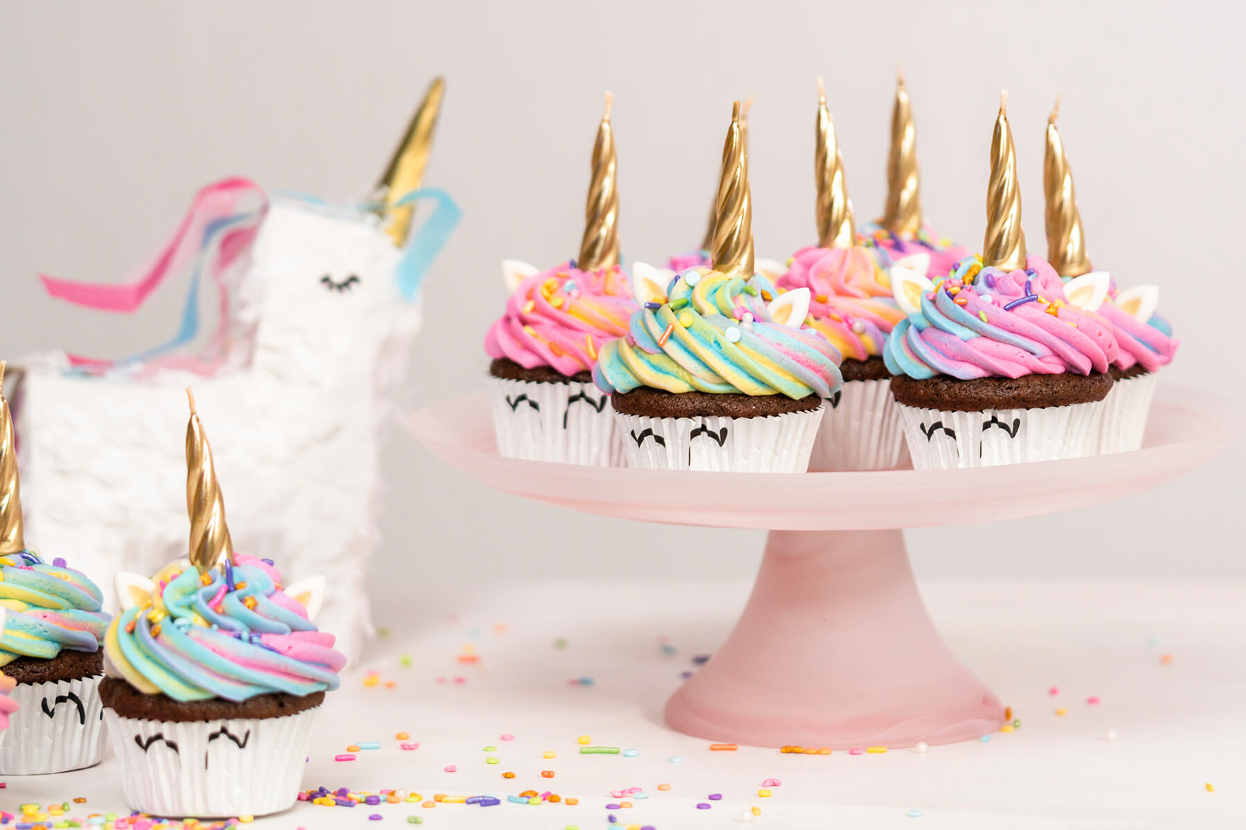 Einhorn-Cupcakes mit goldenen Hörnern auf einem rosa Tortenständer vor einer weißen Einhorn-Torte