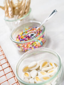 Schälchen mit weißen Fondant-Ohren mit goldenem Innenohr, bunten Zuckerstreuseln und ein Glas mit goldenen Einhorn-Kerzen in Hornform