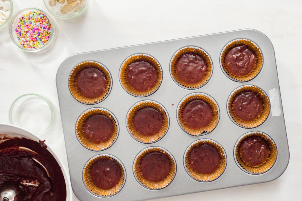 Muffinblech mit zwölf zu zwei Dritteln gefüllten Papierförmchen voll dunklem Schokoladenteig