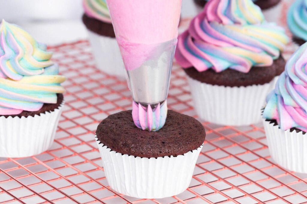 Spritzbeutel mit großer Sterntülle setzt Regenbogen-Buttercreme auf einen ausgekühlten Schokoladen-Cupcake
