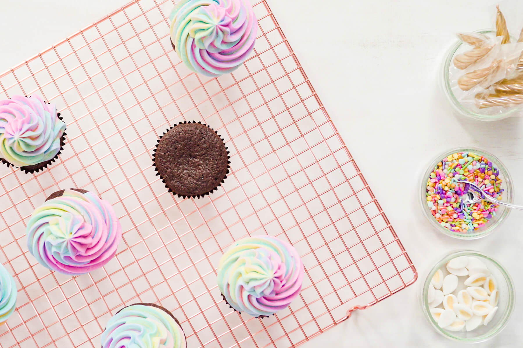Flat-Lay mit gespritzten Regenbogen-Cupcakes auf einem kupferfarbenen Kuchengitter, einem noch unverzierten Schokoladen-Cupcake und Schälchen mit Fondant-Ohren, Streuseln und goldenen Einhorn-Kerzen