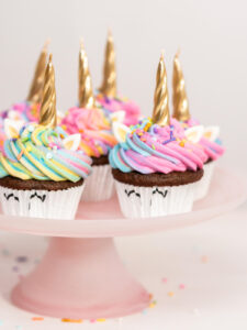 Fertige Einhorn-Cupcakes mit goldenen Hörnern, weißen Ohren und Regenbogenhaube auf einem rosa Tortenständer