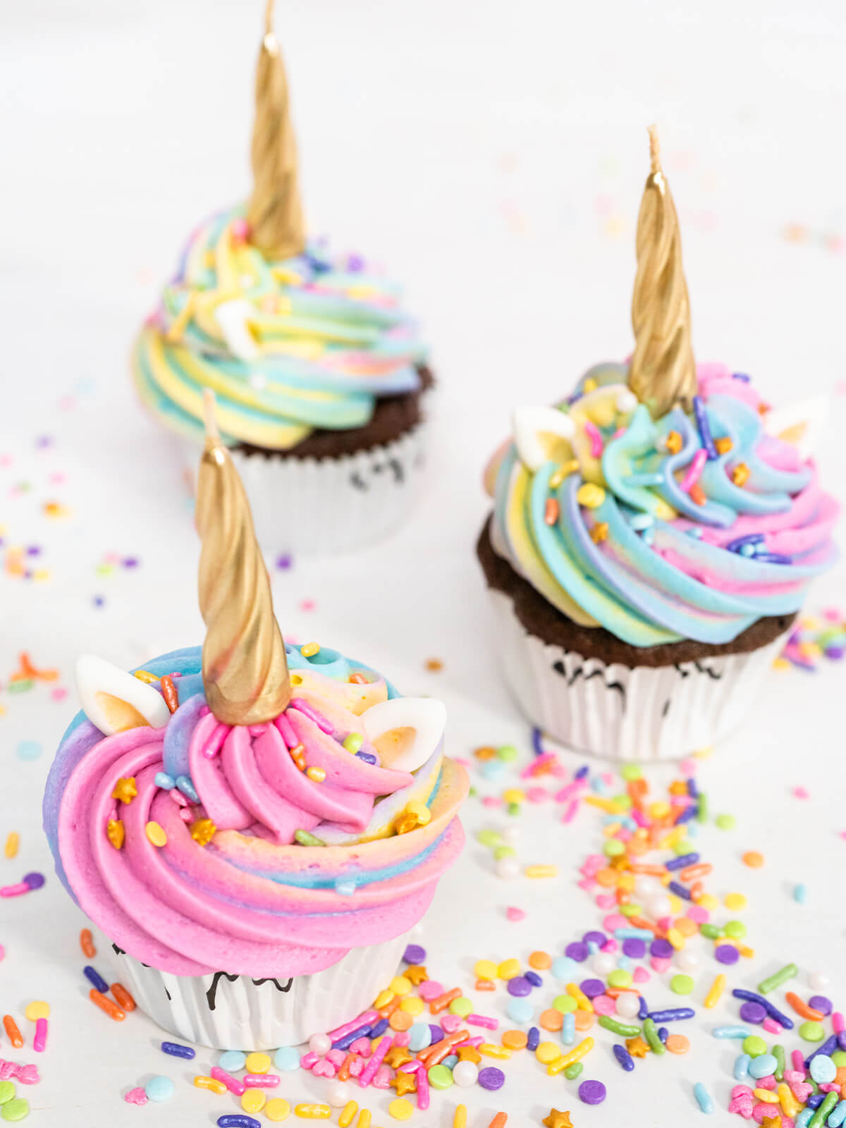 Drei Einhorn-Cupcakes mit goldenen Hörnern, weißen Ohren und Regenbogen-Buttercreme zwischen verstreuten Zuckerstreuseln
