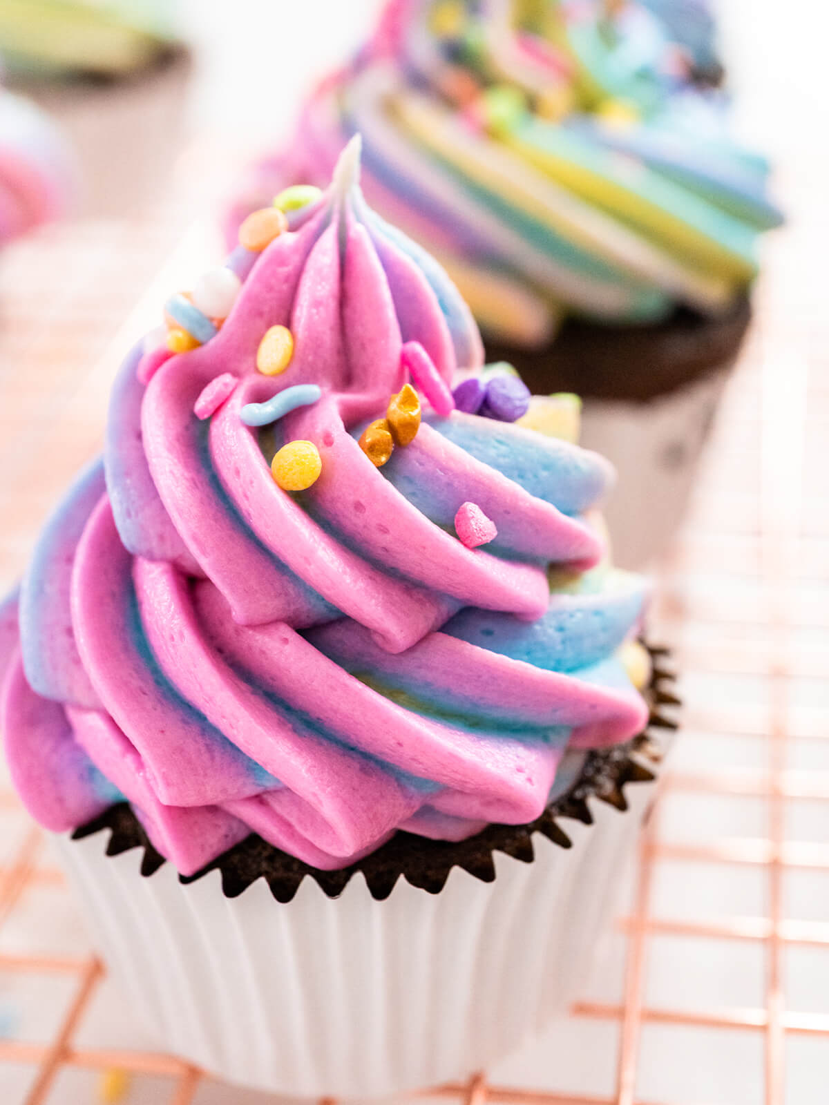 Nahaufnahme einer rosa-blauen Regenbogen-Buttercremehaube mit pastellfarbenen Streuseln auf einem Schokoladen-Cupcake