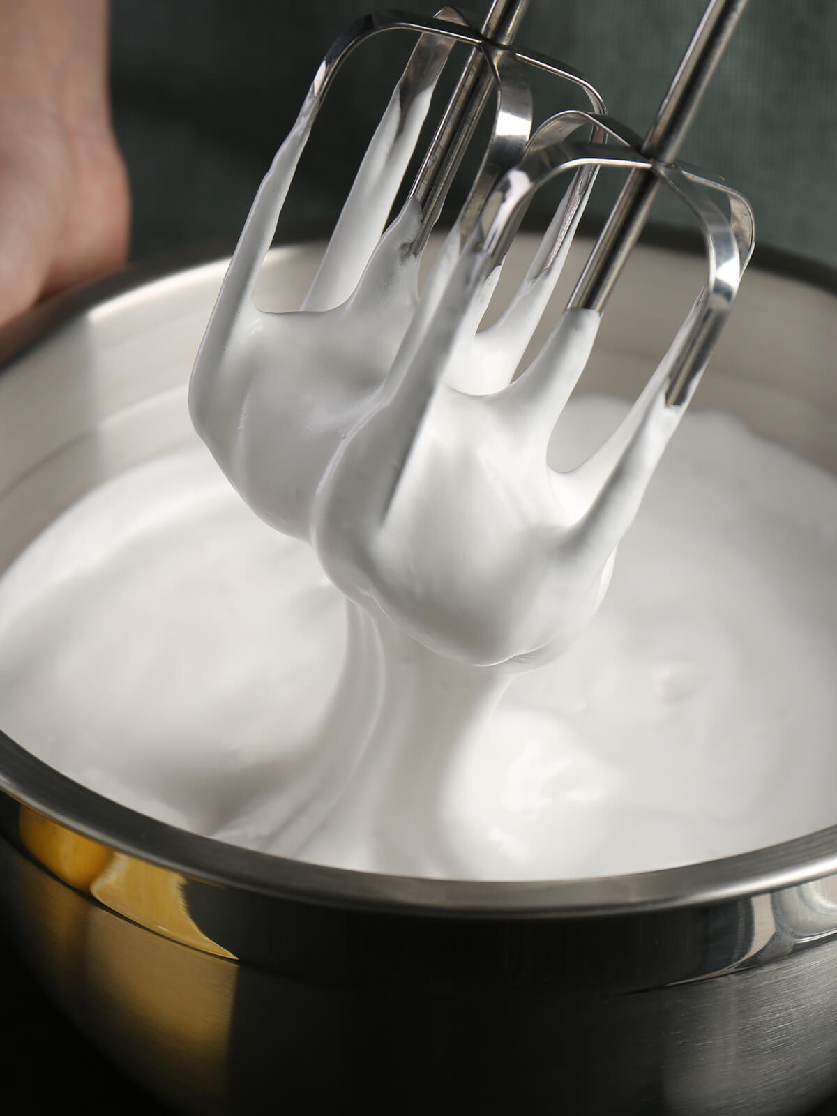 Angehobene Quirle eines Handmixers über einer Edelstahlschüssel, dicht mit weißer, aufgeschlagener Eiweißmasse belegt