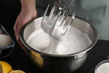 Handmixer mit aufgeschlagenem Eiweiß über einer Edelstahlschüssel, daneben Eier, halbierte Zitronen und eine Schale Zucker