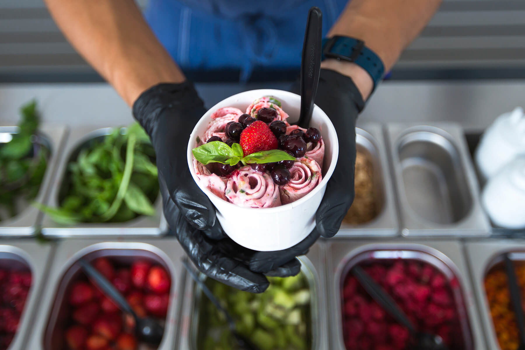 Blick von oben auf einen Becher mit Eisrollen, Kirschen, Erdbeere und Basilikum über der Theke mit Beeren, Kiwi und Kräutern