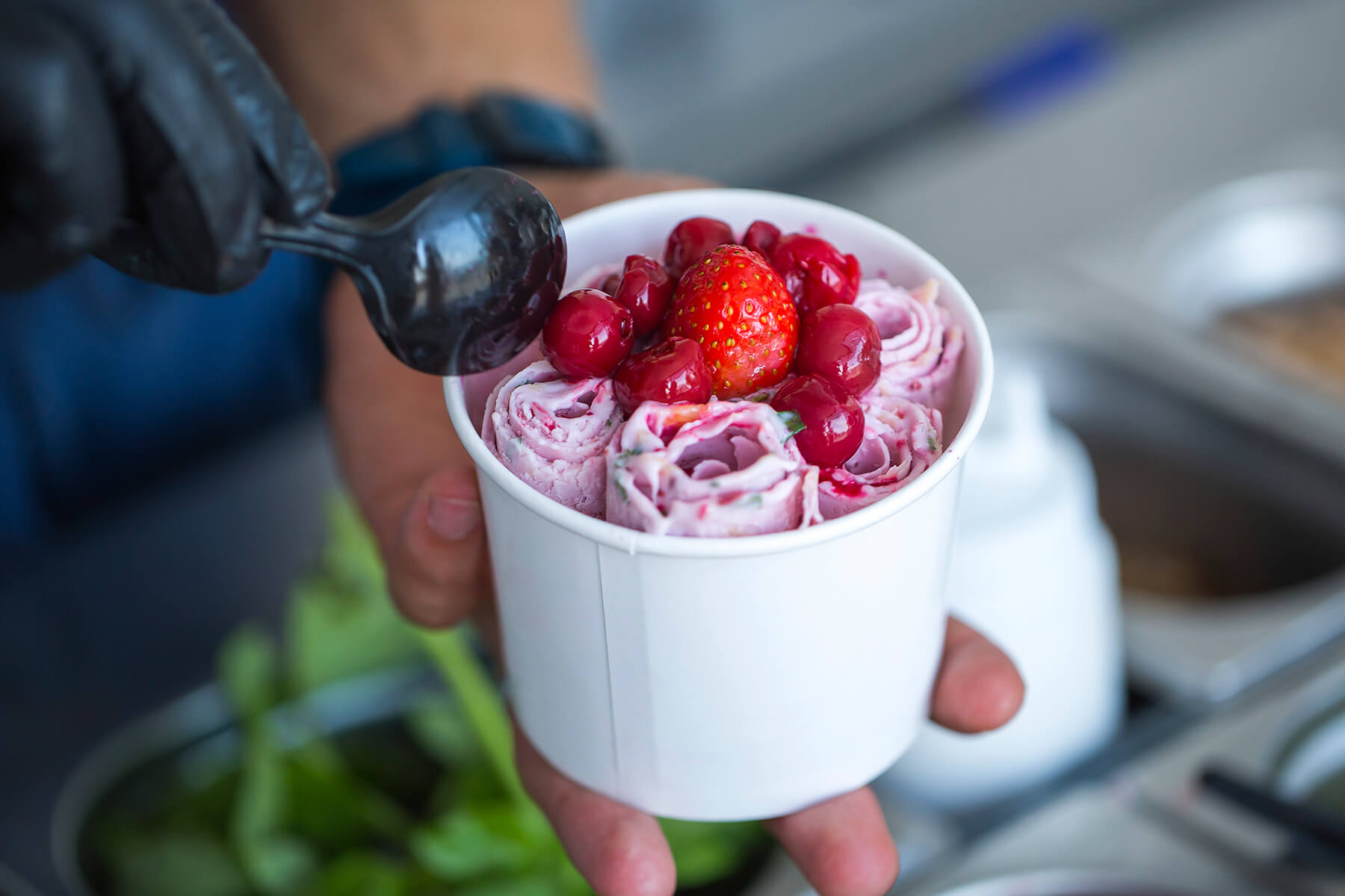 Aus einem Löffel läuft dunkle Kirschsauce über die aufrecht stehenden Eisrollen im Becher, daneben eine frische Erdbeere