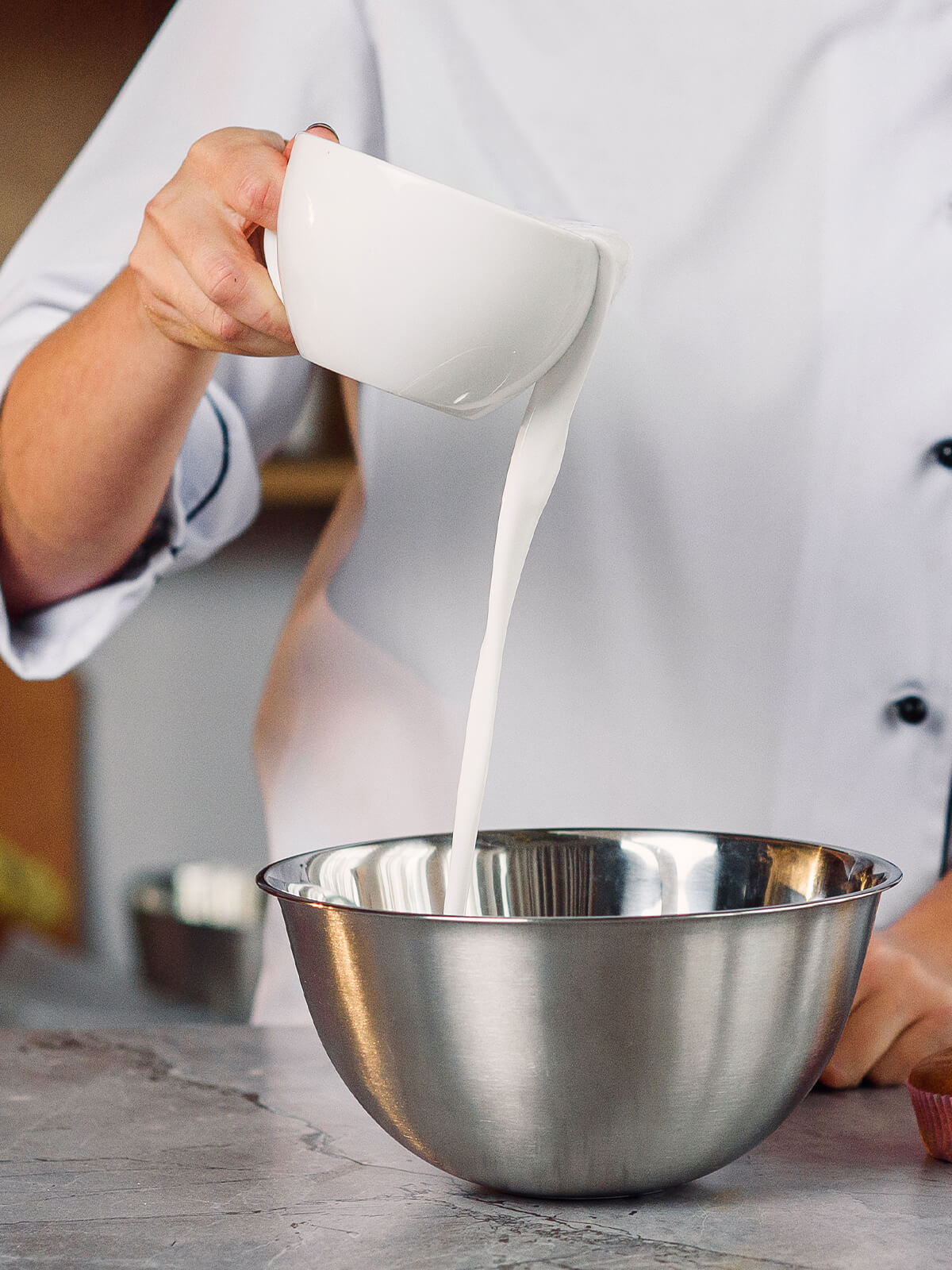 Eine weiße Flüssigkeit wird aus einer Tasse in eine Rührschüssel aus Edelstahl gegossen