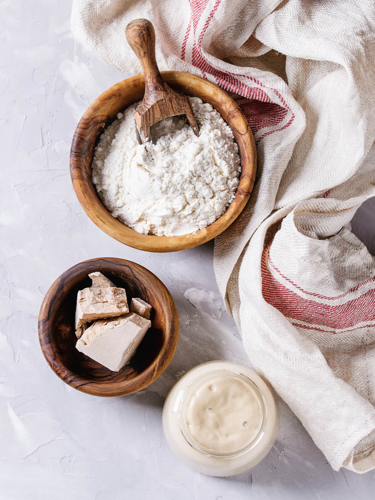 Schale mit Mehl und Holzschaufel, Schale mit gebrochener frischer Hefe und ein Glas Anstellgut auf hellem Untergrund