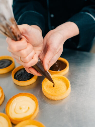 Aus einem Spritzbeutel läuft dunkle Ganache in einen leeren gebackenen Tarteletteboden, daneben stehen gefüllte Böden