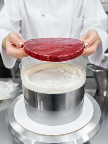 Eine gefrorene rote Fruchtscheibe wird über eine helle Cremeschicht in einem Tortenring gehalten