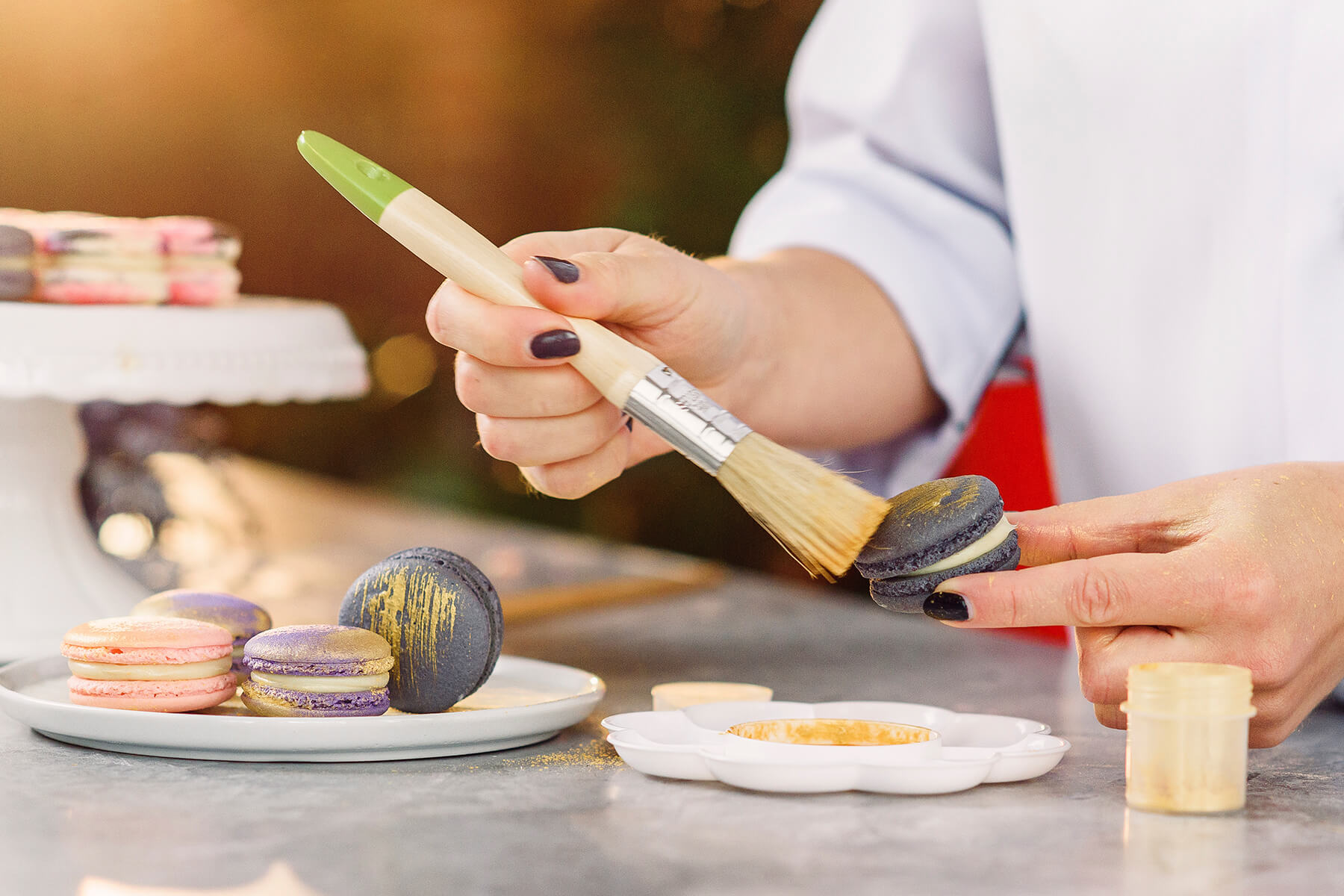 Ein breiter Naturborstenpinsel streicht Goldpuder über ein dunkles Macaron, auf dem Teller daneben liegen Macarons mit unterschiedlich starkem Goldauftrag
