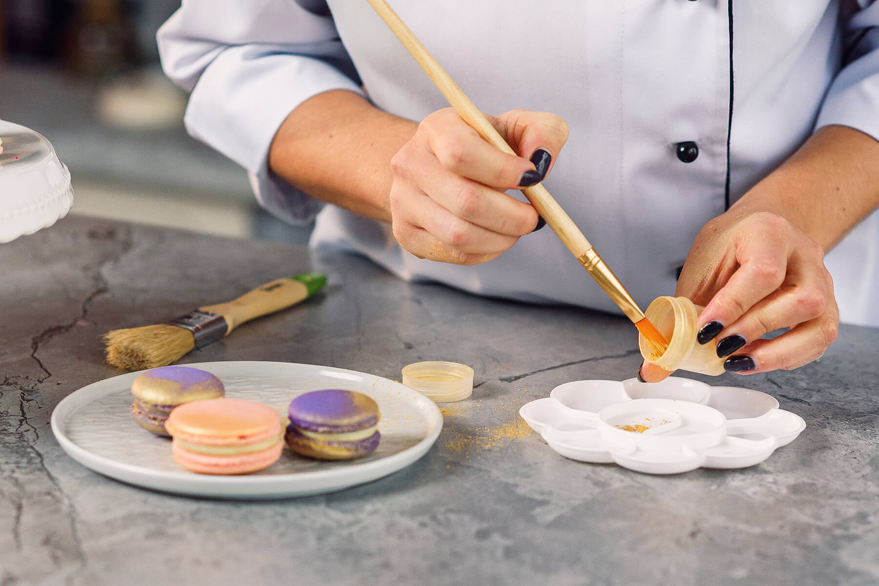 Auf der Arbeitsfläche liegen ein breiter Backpinsel und ein schmaler Flachpinsel, daneben eine Blütenpalette und ein Töpfchen mit Goldpuder