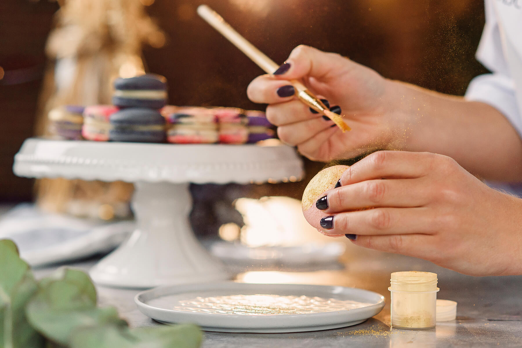 Goldpuder stäubt in einer feinen Wolke durch die Luft, während ein Macaron über einem Teller vergoldet wird, auf dem sich loser Puder sammelt