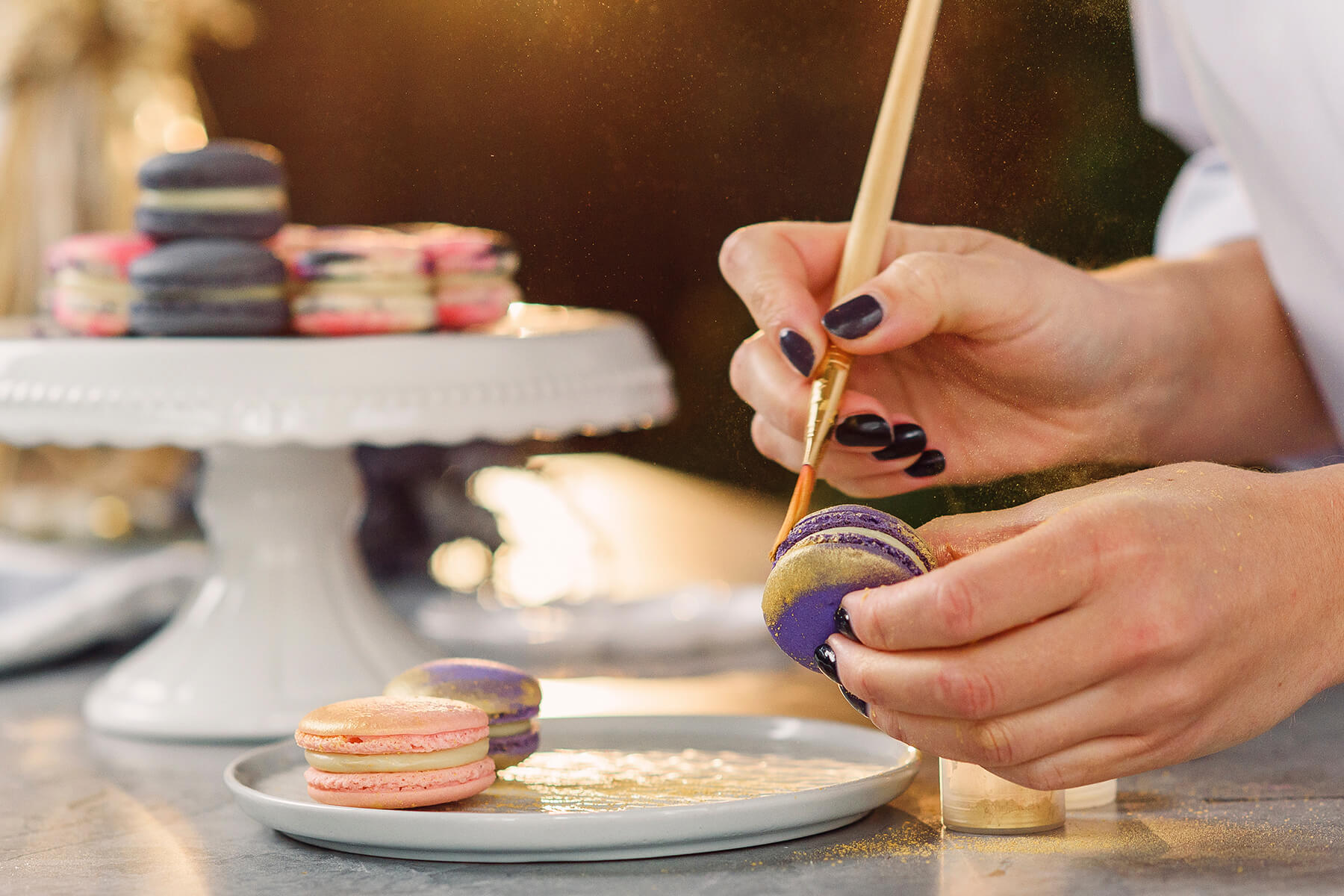 Ein feiner Pinsel zieht einen scharf begrenzten Goldstreifen über den Rand eines violetten Macarons