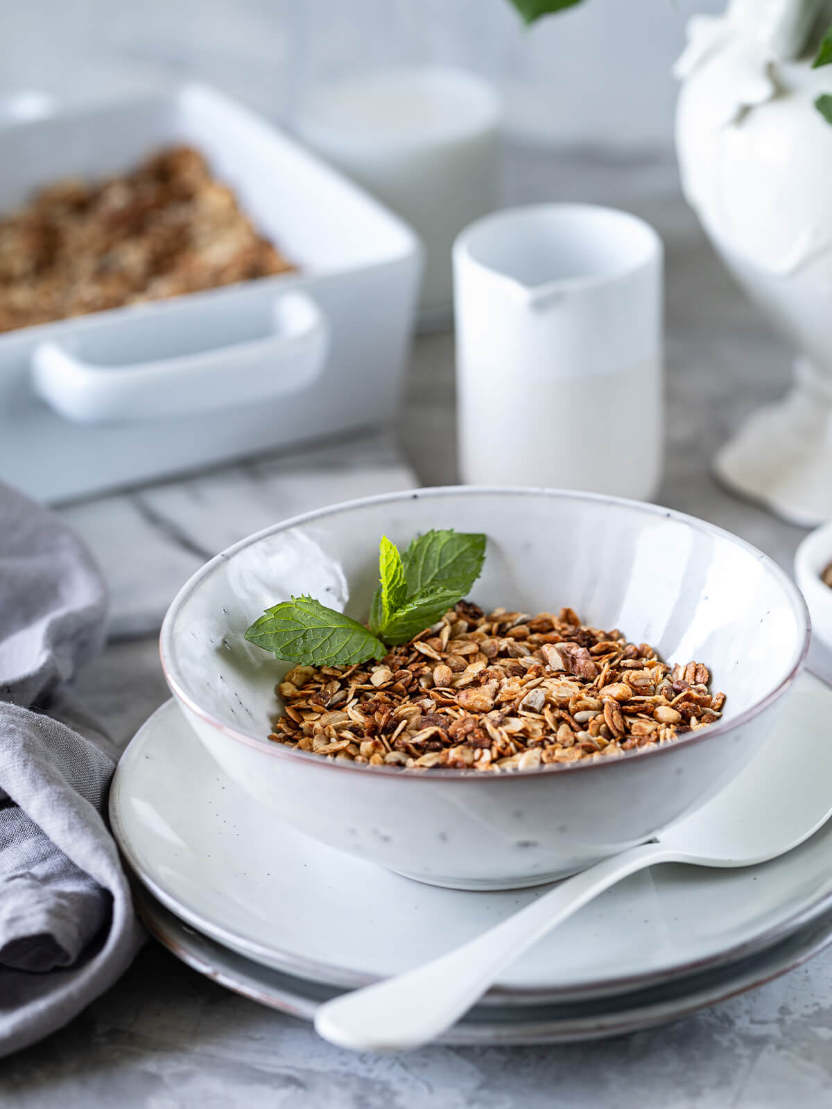 Portion gebackenes Granola in einer weißen Schale mit einem Minzezweig, dahinter eine Auflaufform und ein Milchkännchen