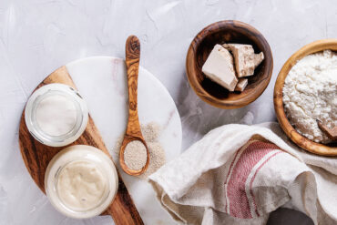 Frische Hefe im Würfel, Trockenhefe-Granulat auf einem Holzlöffel, zwei Gläser Anstellgut und eine Schale Mehl von oben
