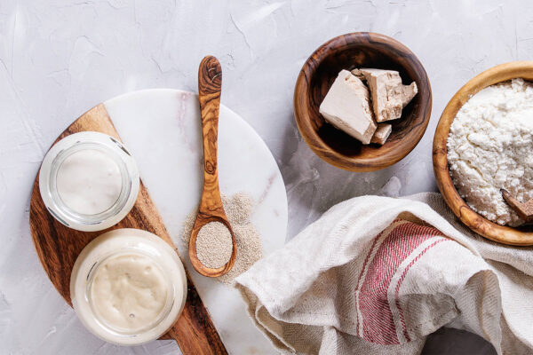 Frische Hefe im Würfel, Trockenhefe-Granulat auf einem Holzlöffel, zwei Gläser Anstellgut und eine Schale Mehl von oben
