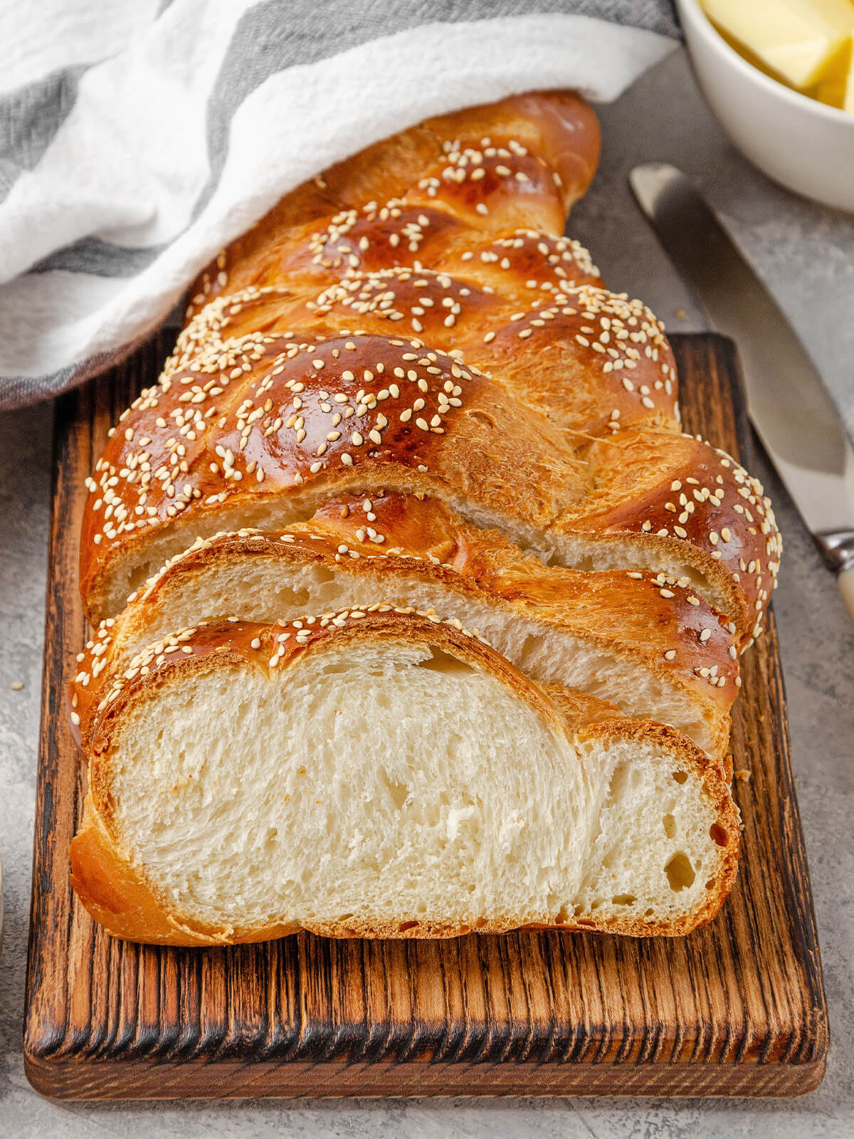 Angeschnittener Hefezopf mit Sesam auf einem dunklen Holzbrett, drei Scheiben zeigen die feine weiße Krume, daneben Butter und ein Messer
