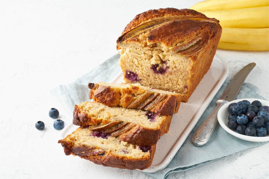 Angeschnittenes Bananenbrot mit Heidelbeeren und Bananenhälften auf einer weißen Platte