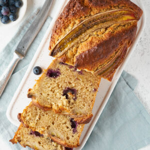 Angeschnittenes Bananenbrot mit Heidelbeeren von oben auf einer weißen Platte, daneben eine Schale Heidelbeeren