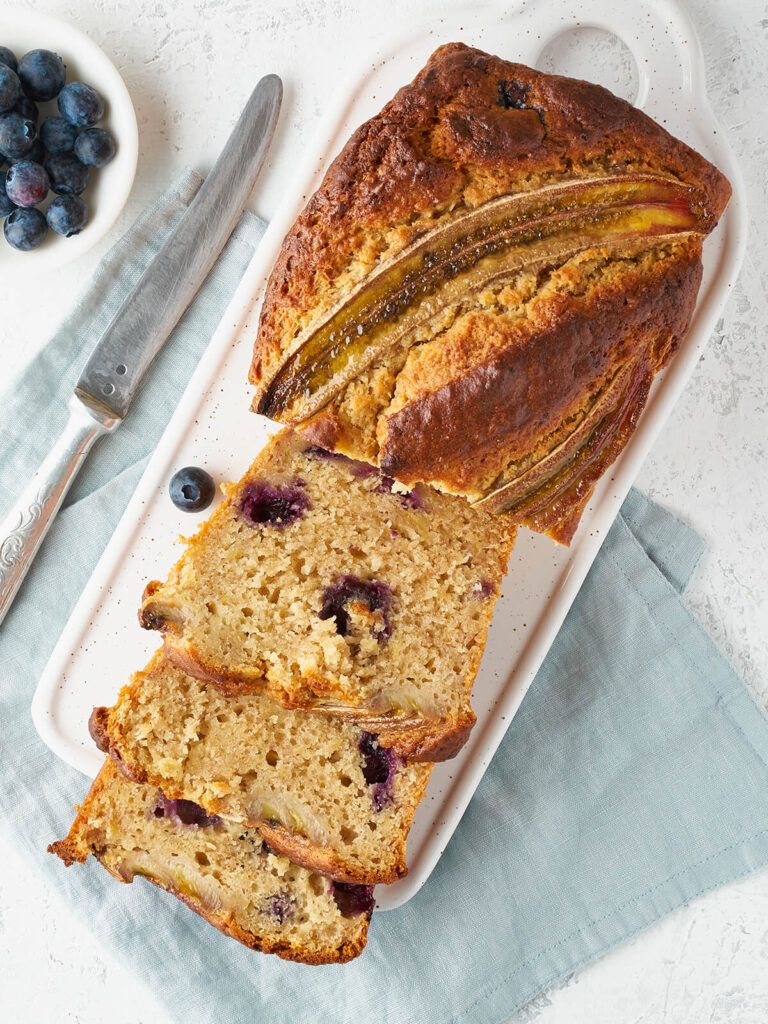 Angeschnittenes Bananenbrot mit Heidelbeeren von oben auf einer weißen Platte, daneben eine Schale Heidelbeeren
