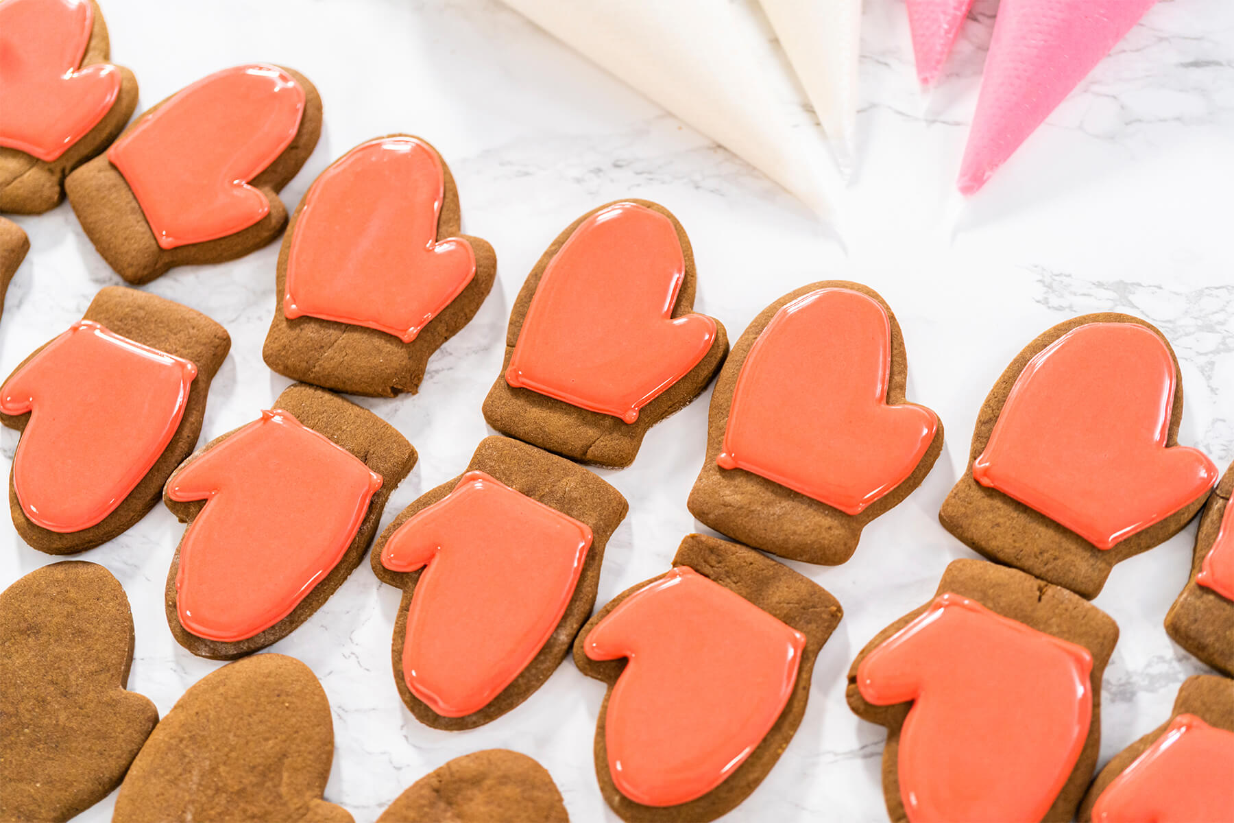 Rot ausgefüllte Lebkuchen-Fäustlinge mit unglasierter Manschette neben gefüllten Spritzbeuteln mit weißer und rosa Glasur