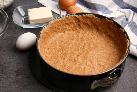 Springform mit fertig angedrücktem Keksboden und hochgezogenem Rand, daneben Butter im Glasschälchen und Eier