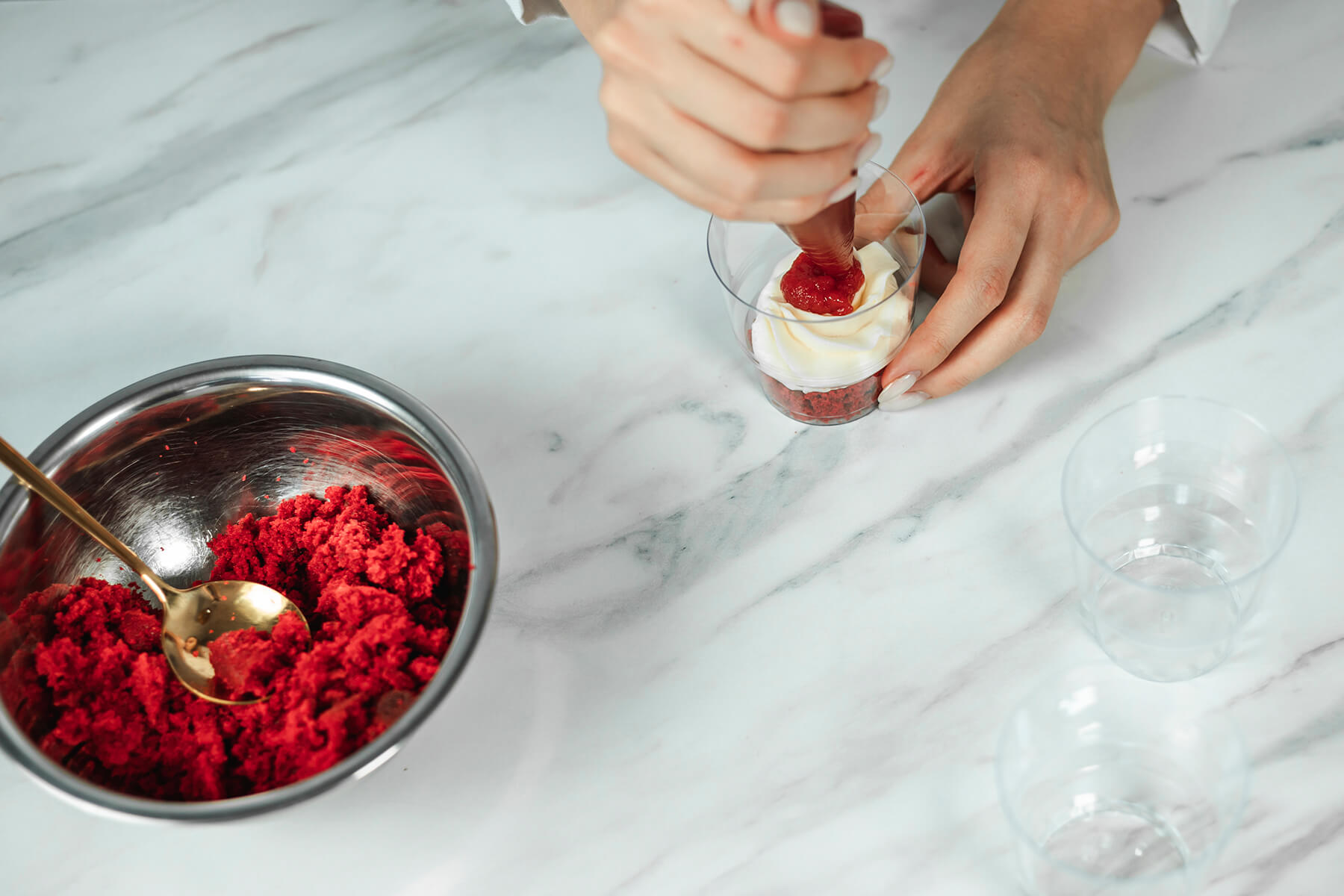 Kuchenkrümel und weiße Creme werden in einem Dessertglas geschichtet, daneben eine Schüssel mit roten Krümeln