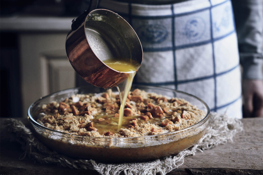 Zerlassene Butter wird aus einem Kupfertopf in die Mitte der Krümelschicht gegossen