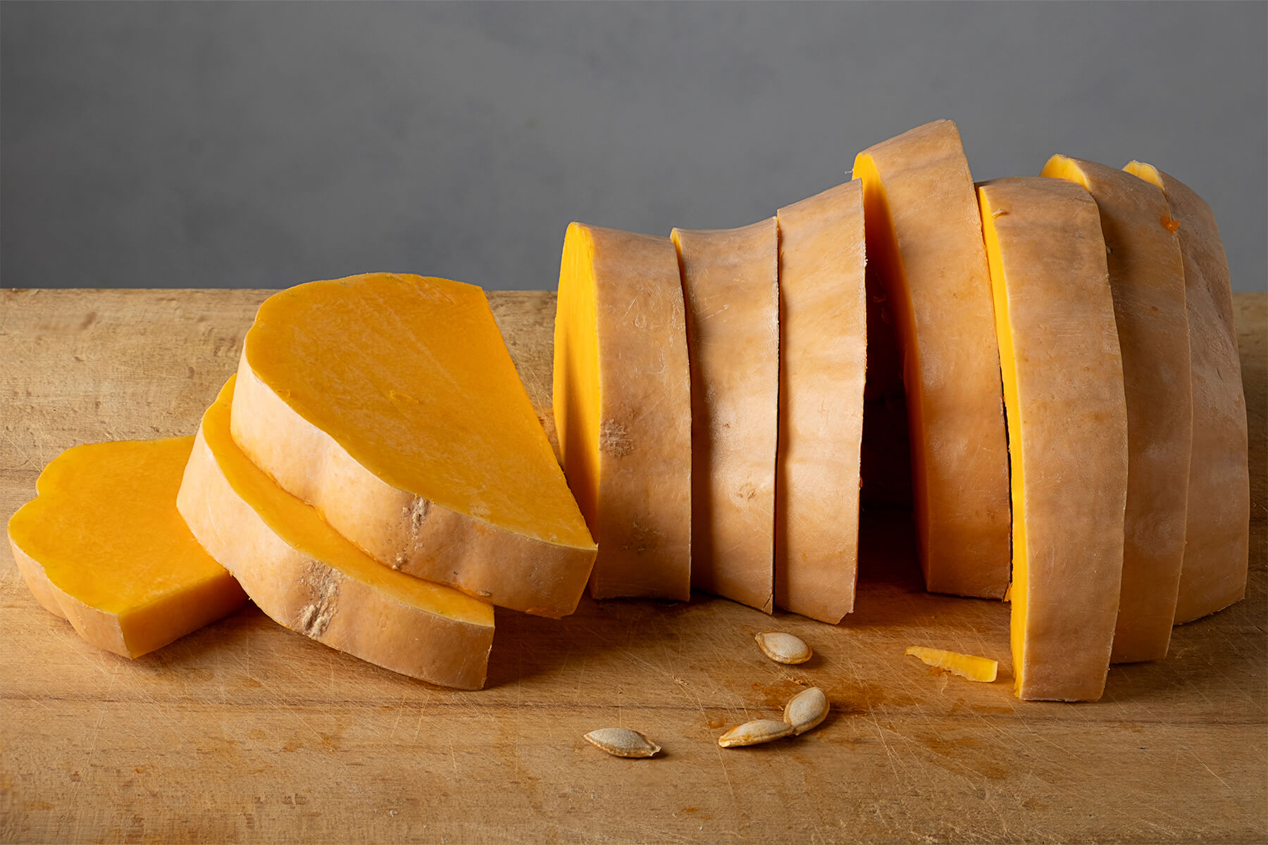 Ein Butternut-Kürbishals ist in etwa zwei Zentimeter dicke Scheiben geschnitten und liegt auf einem Holzbrett, davor liegen einzelne helle Kürbiskerne