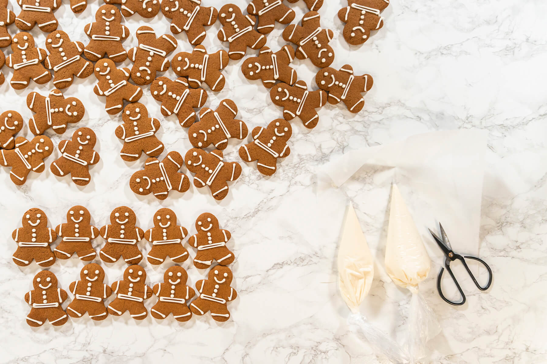 Ausgekühlte Lebkuchen-Männchen auf Marmor, daneben zwei gefüllte Spritzbeutel mit Eiweißglasur und eine Schere