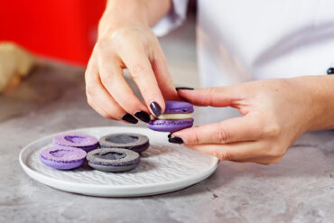 Vier leere Macaronschalen in Violett und Grau liegen mit der Unterseite nach oben, jede mit einem eingesunkenen Krater in der Mitte
