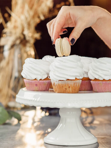 Zwei Finger setzen ein Macaron aufrecht auf die weiße Cremehaube eines Cupcakes