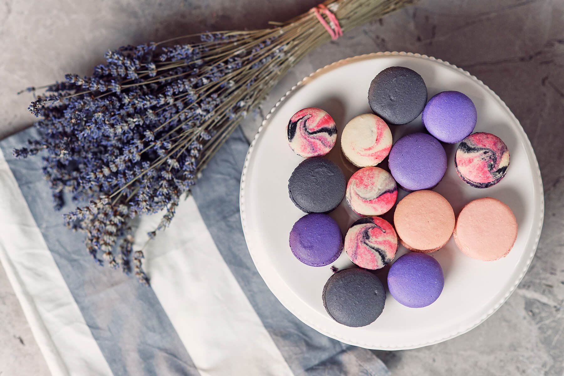 Vierzehn Macarons in Anthrazit, Violett, Rosa und mit pink-schwarzer Marmorierung liegen dicht an dicht auf einer weißen Tortenplatte