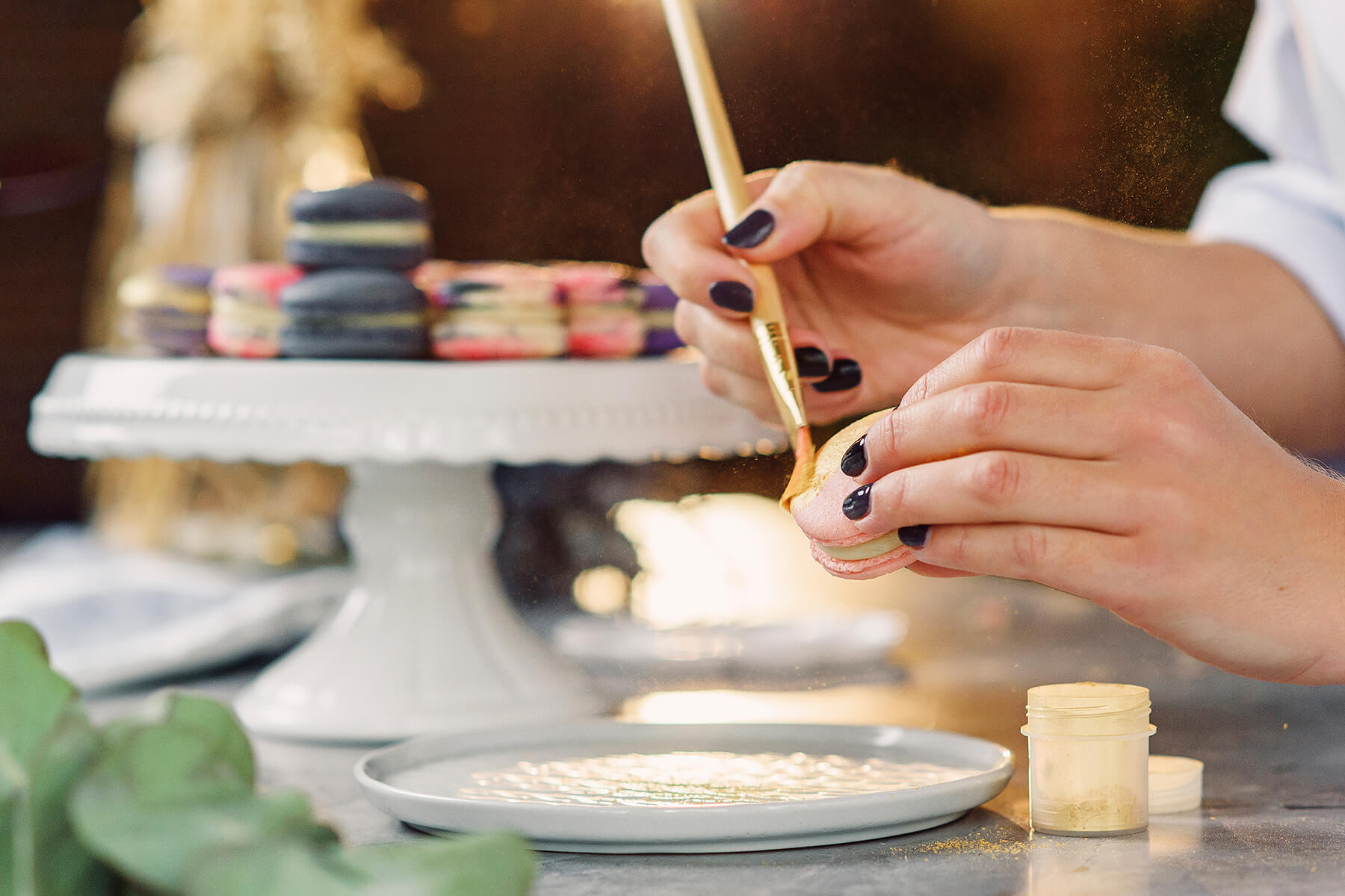 Eine Etagere mit anthrazitfarbenen, violetten und rosa marmorierten Macarons, im Vordergrund ein Stück beim Vergolden