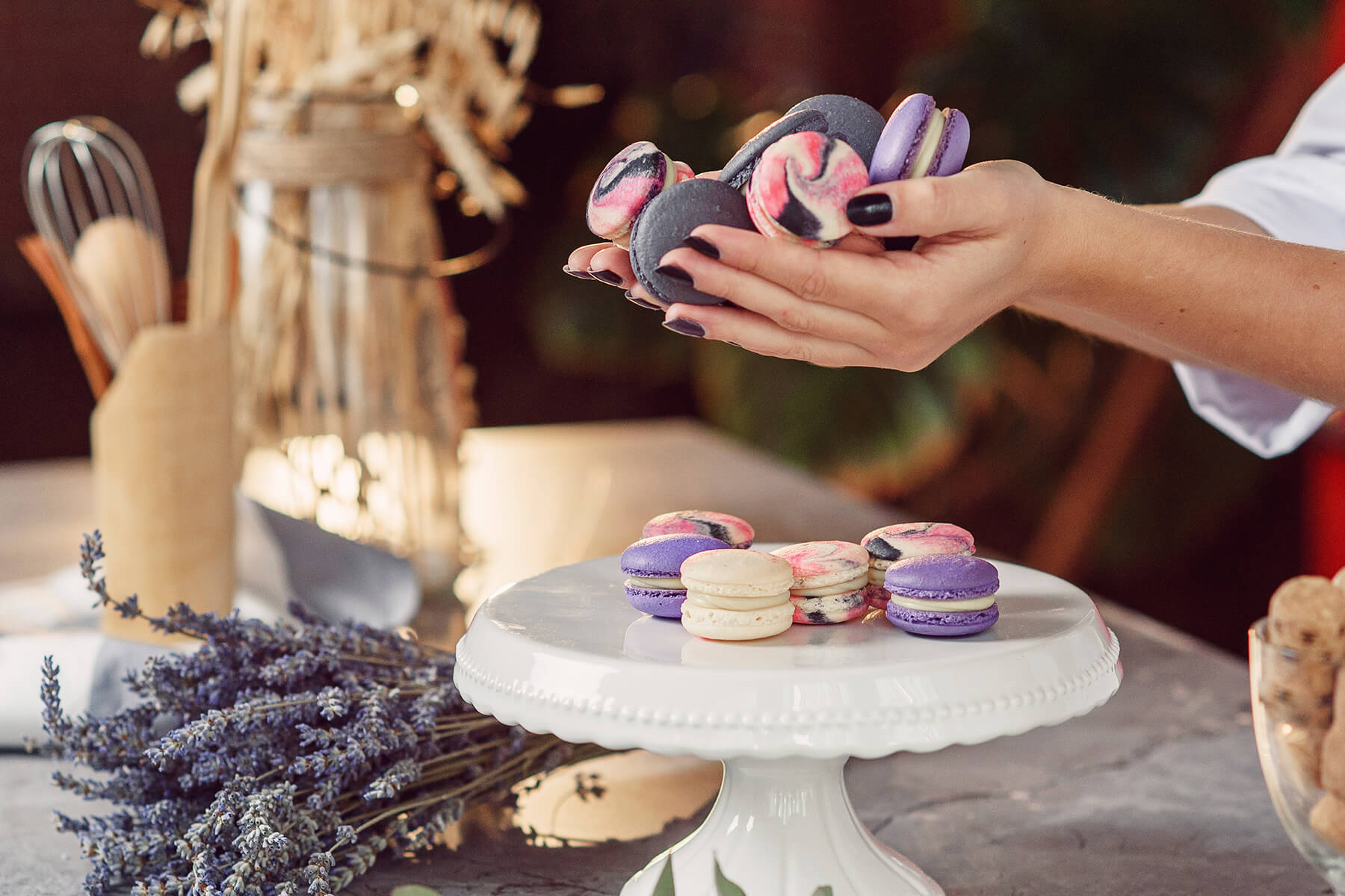 Eine Hand hält marmorierte und violette Macarons über eine Tortenplatte, auf der ein cremefarbenes ungefärbtes Stück neben gefärbten liegt