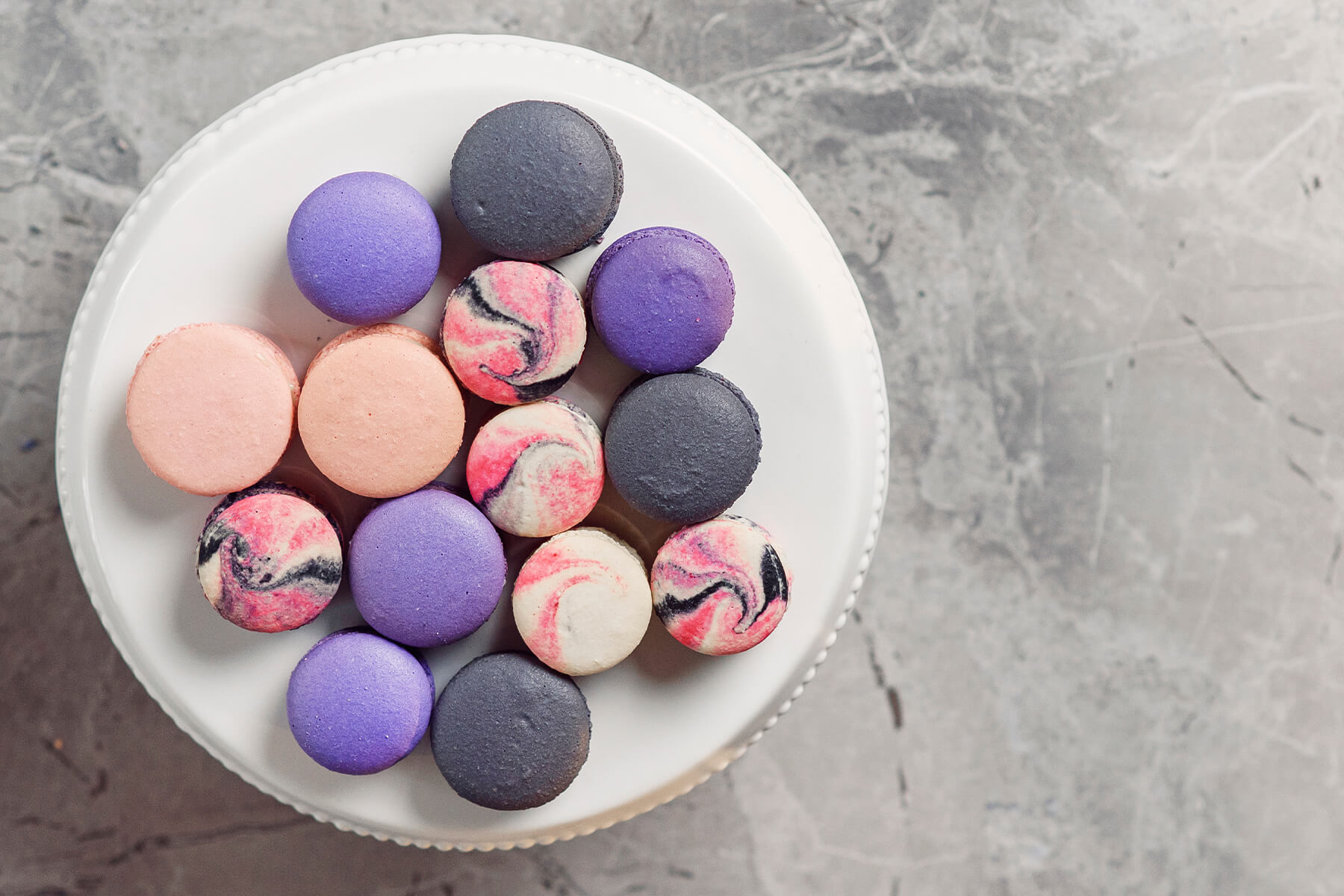 Aufsicht auf eine weiße Platte mit fünf marmorierten Macarons in Pink und Schwarz auf cremefarbenem Grund, dazwischen einfarbige in Violett, Anthrazit und Rosa