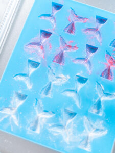 Blaue Silikonform mit rund zwanzig Vertiefungen in Form von Meerjungfrauen-Flossen auf einem Blech, die Vertiefungen sind leer und mit rosa und weißem Perlglanzpuder ausgestäubt, der Puder liegt in den Rillen des Schuppenreliefs und als feiner Schleier auf dem blauen Silikon