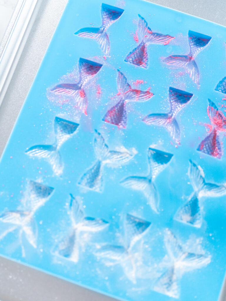 Blaue Silikonform mit rund zwanzig Vertiefungen in Form von Meerjungfrauen-Flossen auf einem Blech, die Vertiefungen sind leer und mit rosa und weißem Perlglanzpuder ausgestäubt, der Puder liegt in den Rillen des Schuppenreliefs und als feiner Schleier auf dem blauen Silikon