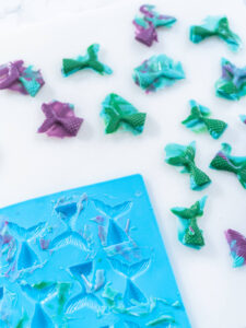 Frisch entformte Flossen in Grün, Türkis und Violett auf heller Fläche, an jeder Flosse steht noch der dünne, unregelmäßige Überstand der Gießmasse ab, unten im Bild die blaue Silikonform mit Resten der erstarrten Masse in und neben den Vertiefungen