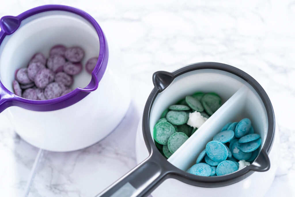 Zwei weiße Schmelztöpfe auf hellem Marmor, im linken violette Schmelzglasur-Drops, im rechten zweigeteilten Topf grüne Drops in der einen und türkisfarbene in der anderen Hälfte, dazwischen jeweils ein kleines Stück festes weißes Kokosfett